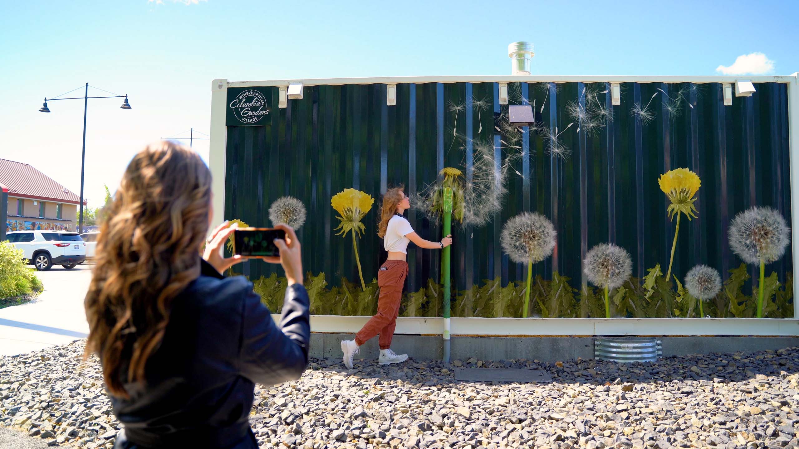 A person snaps a photo of their friend pretending to blow the fluff off a dandelion on the shipping container art wrap at Columbia Gardens Wine & Artisan Village.