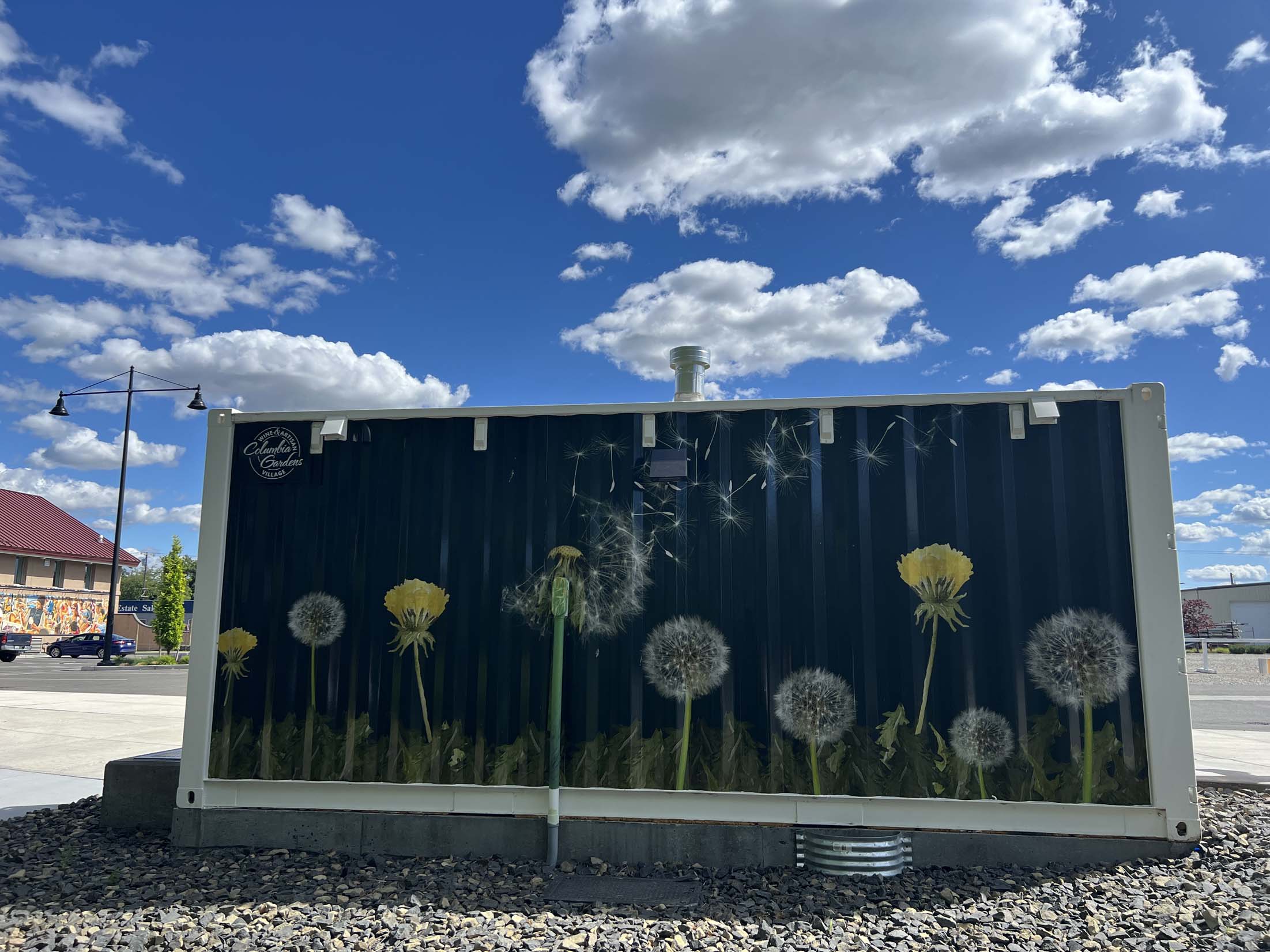 An art wrap with yellow floret and white puffball dandelions fills one side of a shipping container restroom at Columbia Gardens.